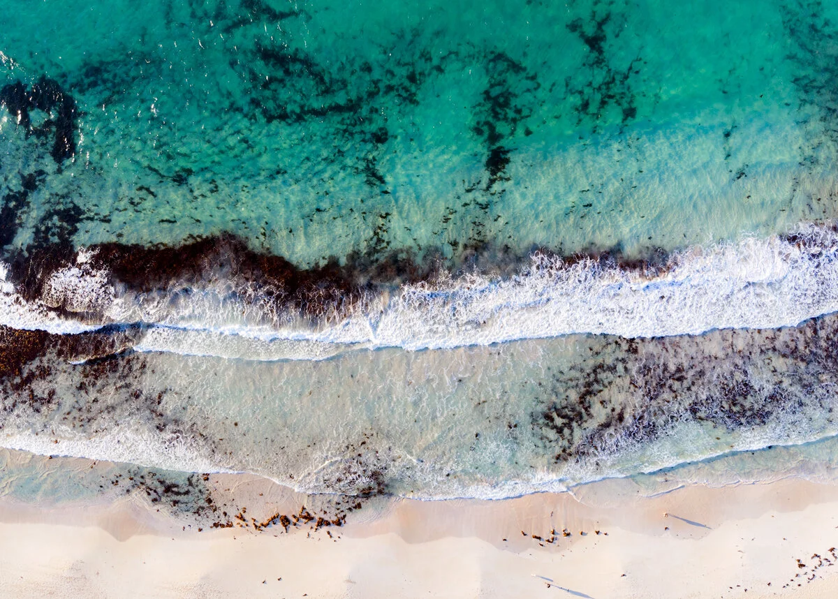 Aerial view of turquoise waves and seaweed patches rolling onto a sandy beach