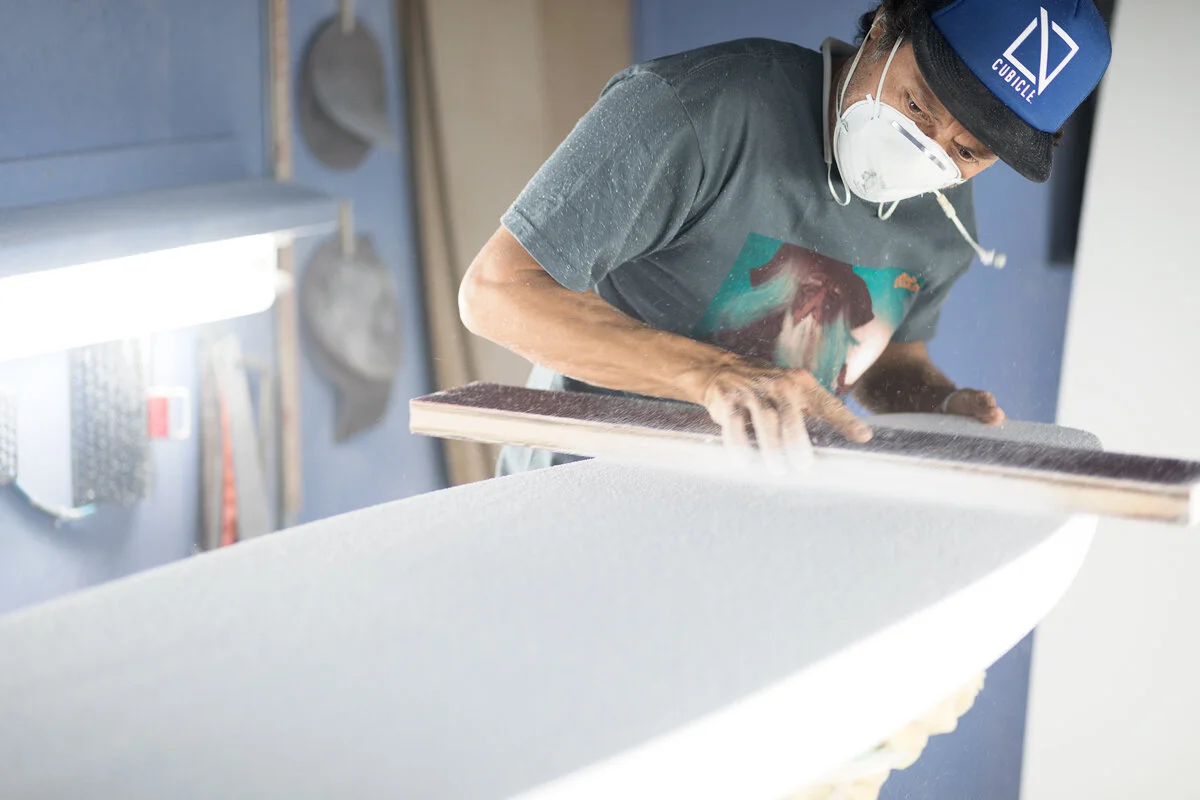 Surfboard shaper sanding a board by hand in his workshop