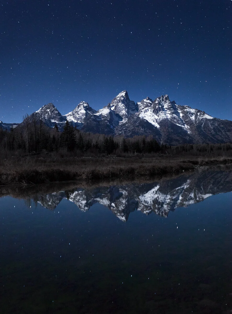 Grand Teton mountains reflected in still water under a starry night sky